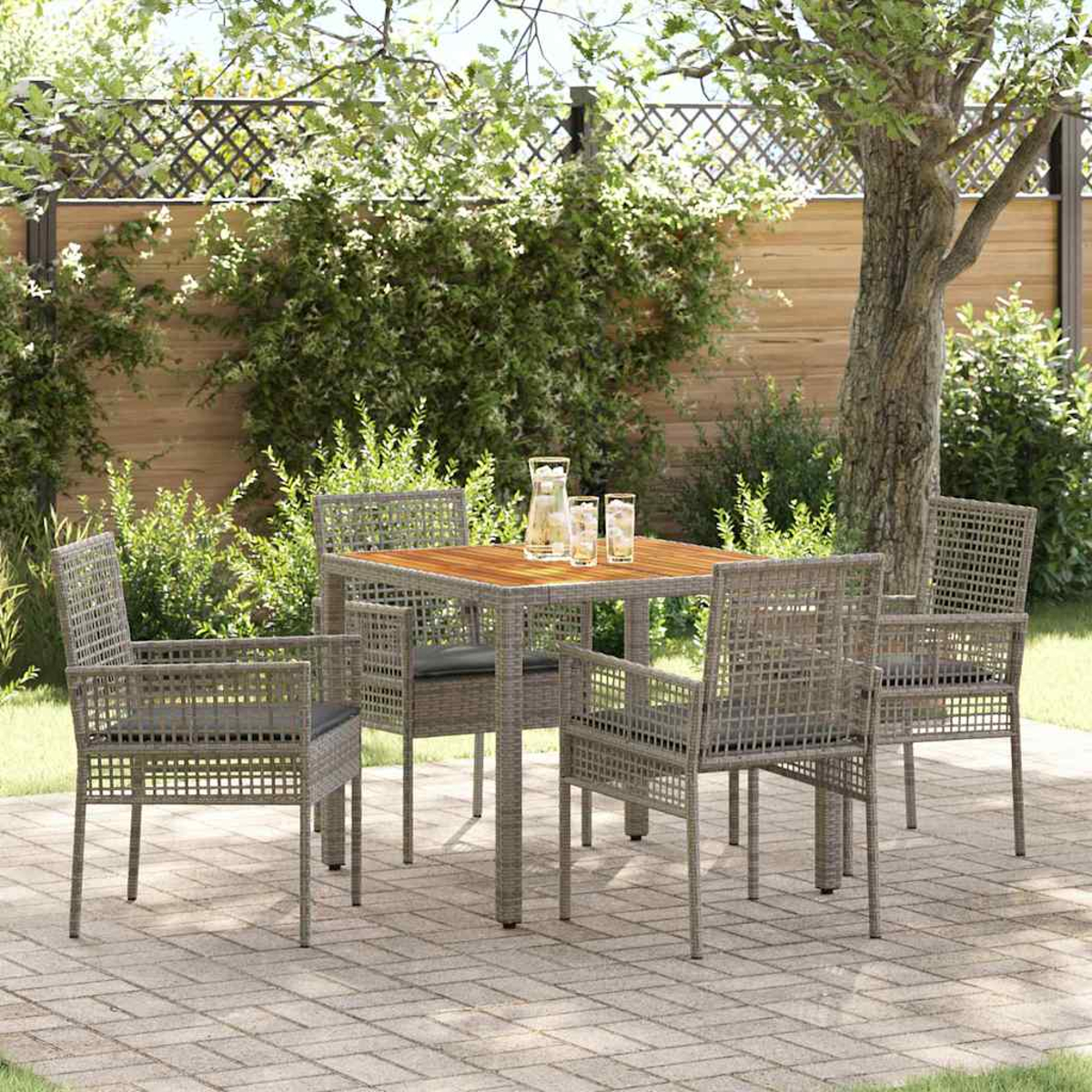 Terrassenset. Quadratischer Holztisch mit vier grauen Korbstühlen. Wasserkrug und Gläser auf dem Tisch. Grünes Laub im Hintergrund.
