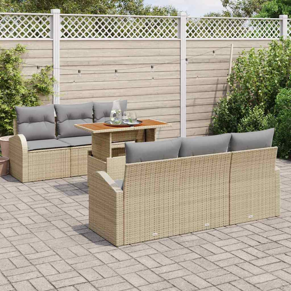 Terrassenset mit beigen Korbmöbeln, grauen Kissen und einem Holztisch. Auf einer gepflasterten Terrasse mit weißem Zaun und Grün.