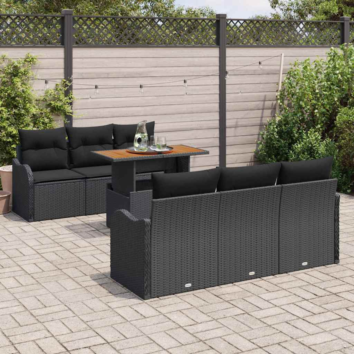 Terrassenmöbel mit schwarzen Kissen, Holztisch und Wasserkrug. Auf Steinfliesen, mit Zaun und Pflanzen im Hintergrund.
