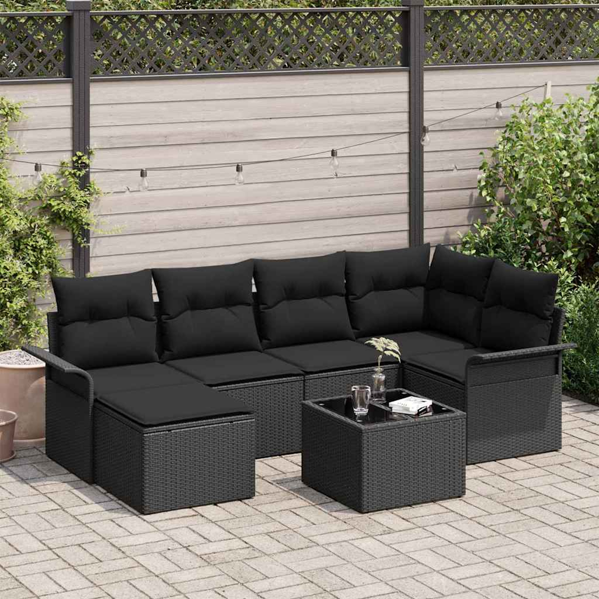 Schwarzes Outdoor-Koriansofa mit Kissen, Glastisch und Lichterkette vor Holzzaun und Grün.