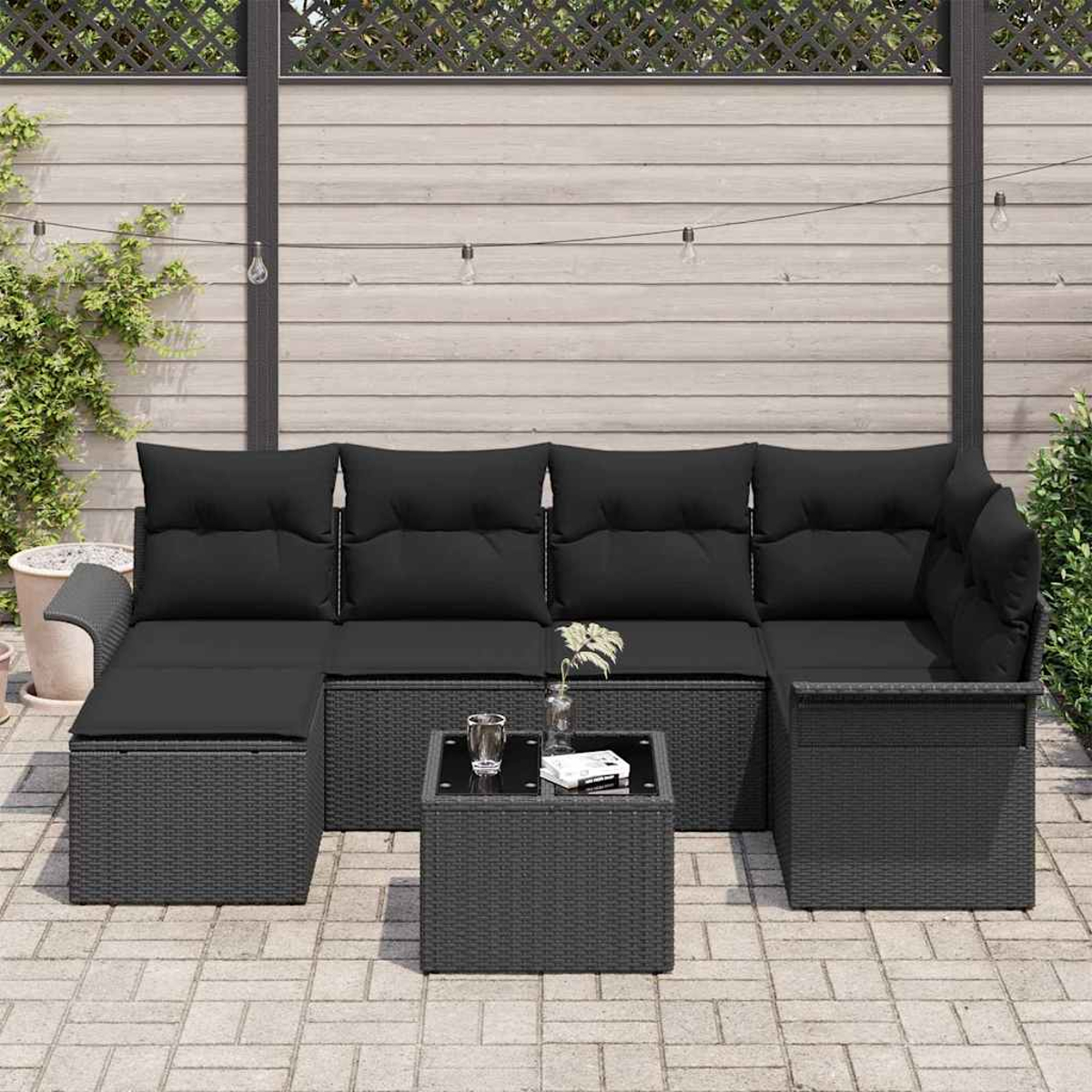 Outdoor-Ecksofa mit schwarzen Kissen und einem kleinen Tisch. Im Hintergrund ein Holzzaun und Pflanzen.