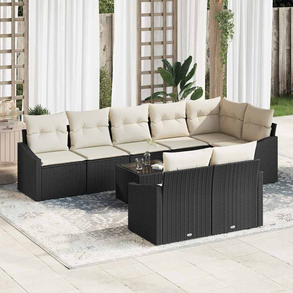 Gartenmöbel-Set: schwarzes Korbmöbel-Sofa mit cremefarbenen Kissen, Tisch mit Glasplatte und Teppich auf einer Terrasse mit Vorhängen.