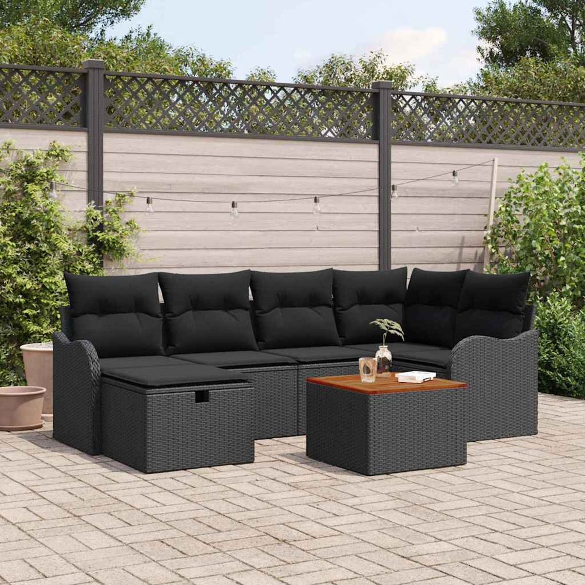 Graues Outdoor-Geflecht-Ecksofa mit schwarzen Kissen und einem Holztisch. Auf einer Ziegelterrasse mit Zaun im Hintergrund.