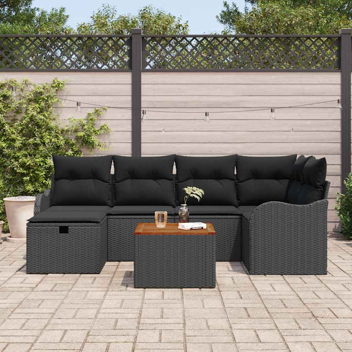 Szene im Freien mit grauem Korbsessel und Couchtisch. Schwarze Kissen. Holzzaun im Hintergrund. Lichterkette. Gepflasterte Terrasse.