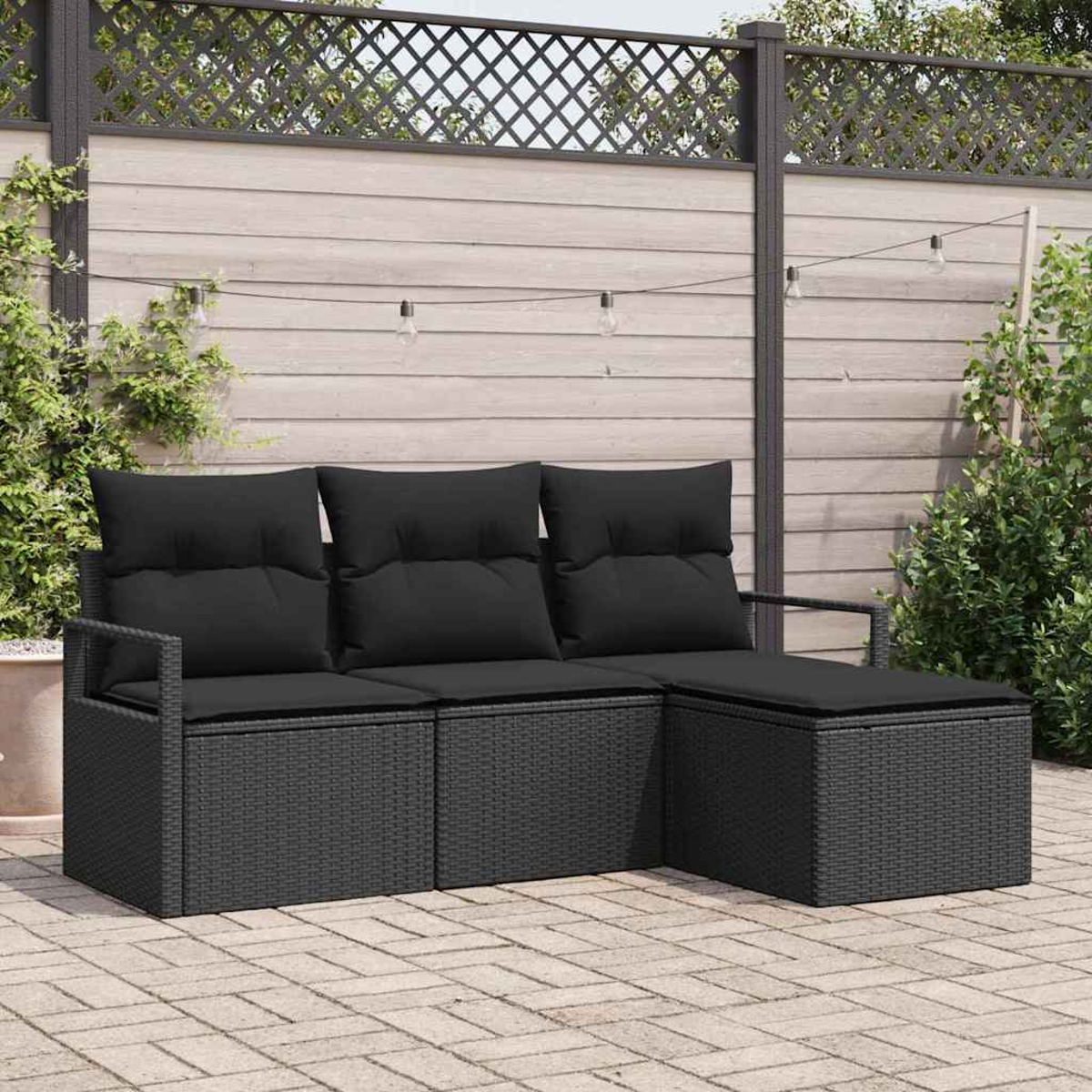 Un sofá seccional de mimbre negro con cojines negros está en un patio. Una valla de madera y luces de cadena están en el fondo.