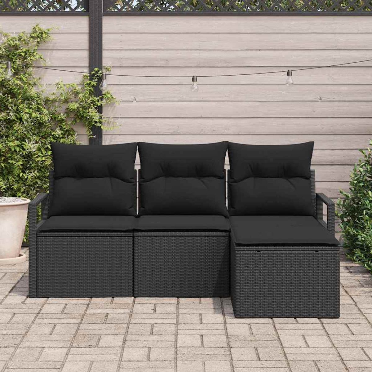 Un sofá seccional de mimbre negro para exteriores con cojines está en un patio pavimentado. Una valla de madera y plantas están al fondo.