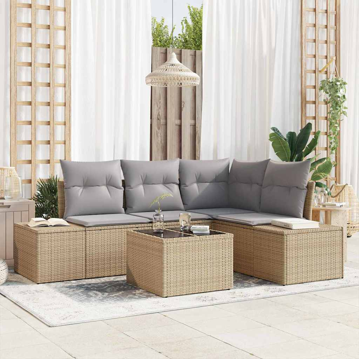 Rattansofa im Freien mit grauen Kissen, Couchtisch mit Glasplatte und weißen Vorhängen. Pflanzen im Hintergrund. Sonniger Terrassenbereich.