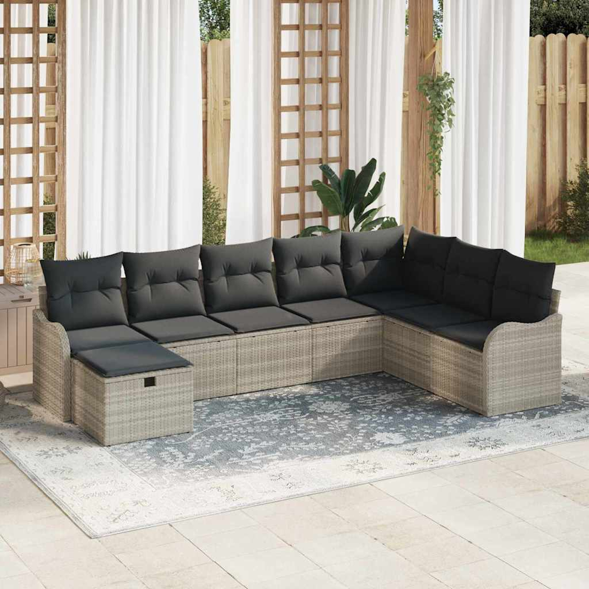 Ein Ecksofa mit schwarzen Kissen steht auf einem gemusterten Teppich. Weiße Vorhänge und Holzkonstruktionen sind im Hintergrund.