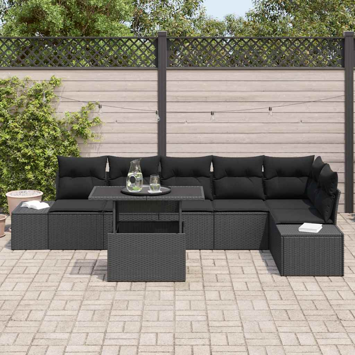 Juego de patio al aire libre. Seccional negro con cojines, mesa con bebidas y un fondo de valla de madera y vegetación.