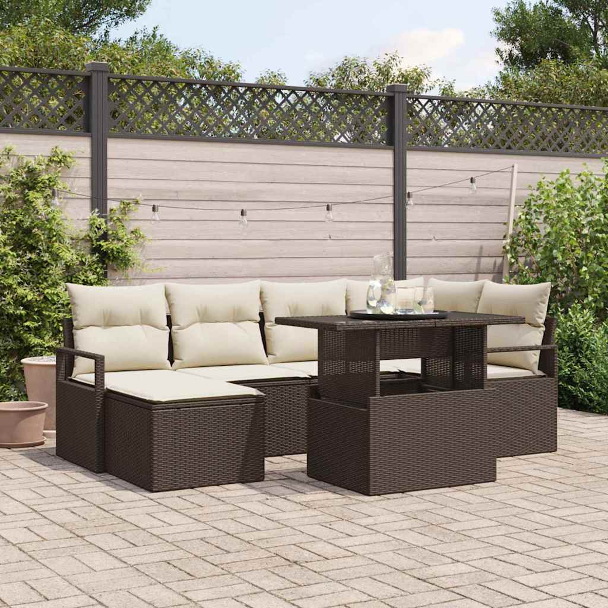 Outdoor-Sofa-Set aus braunem Korbgeflecht mit beigen Kissen und einem passenden Tisch auf einer Terrasse. Im Hintergrund ein Zaun und Grün.
