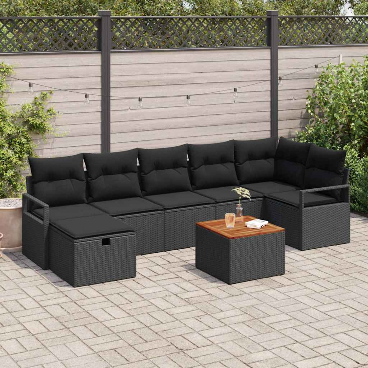 Outdoor-Ecksofa mit schwarzen Kissen und einem kleinen Tisch, auf einer gepflasterten Terrasse. Im Hintergrund ein Holzzaun und Grün.