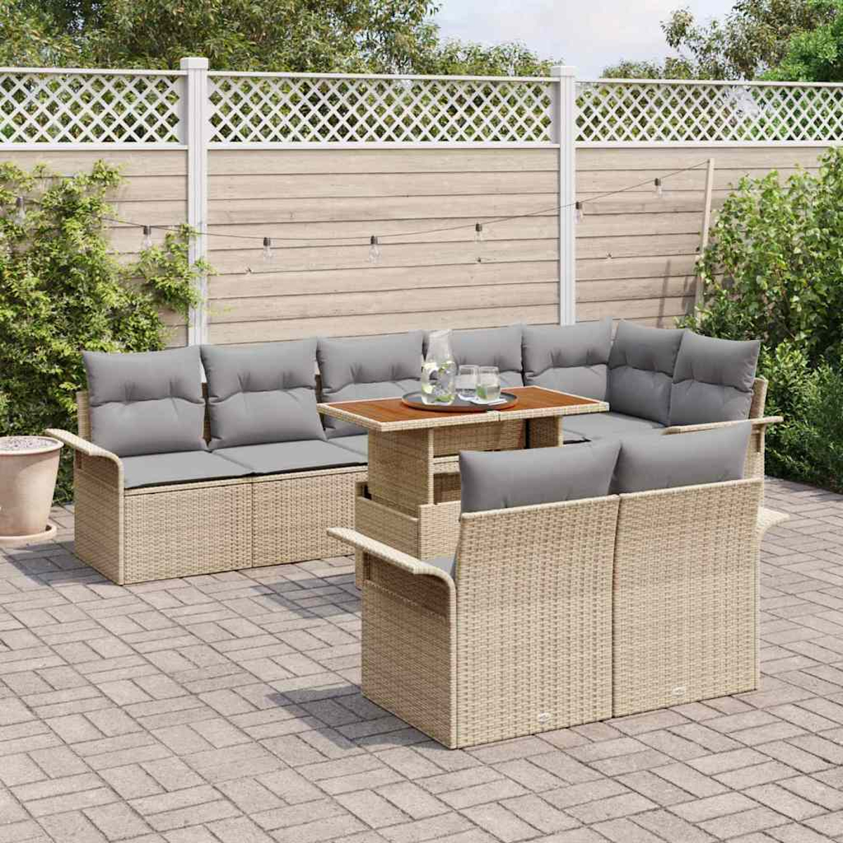 Gartenmöbel-Set aus Korbgeflecht mit grauen Kissen, einem Holztisch mit Getränken, auf einer Backstein-Terrasse, mit Zaun und Grün im Hintergrund.