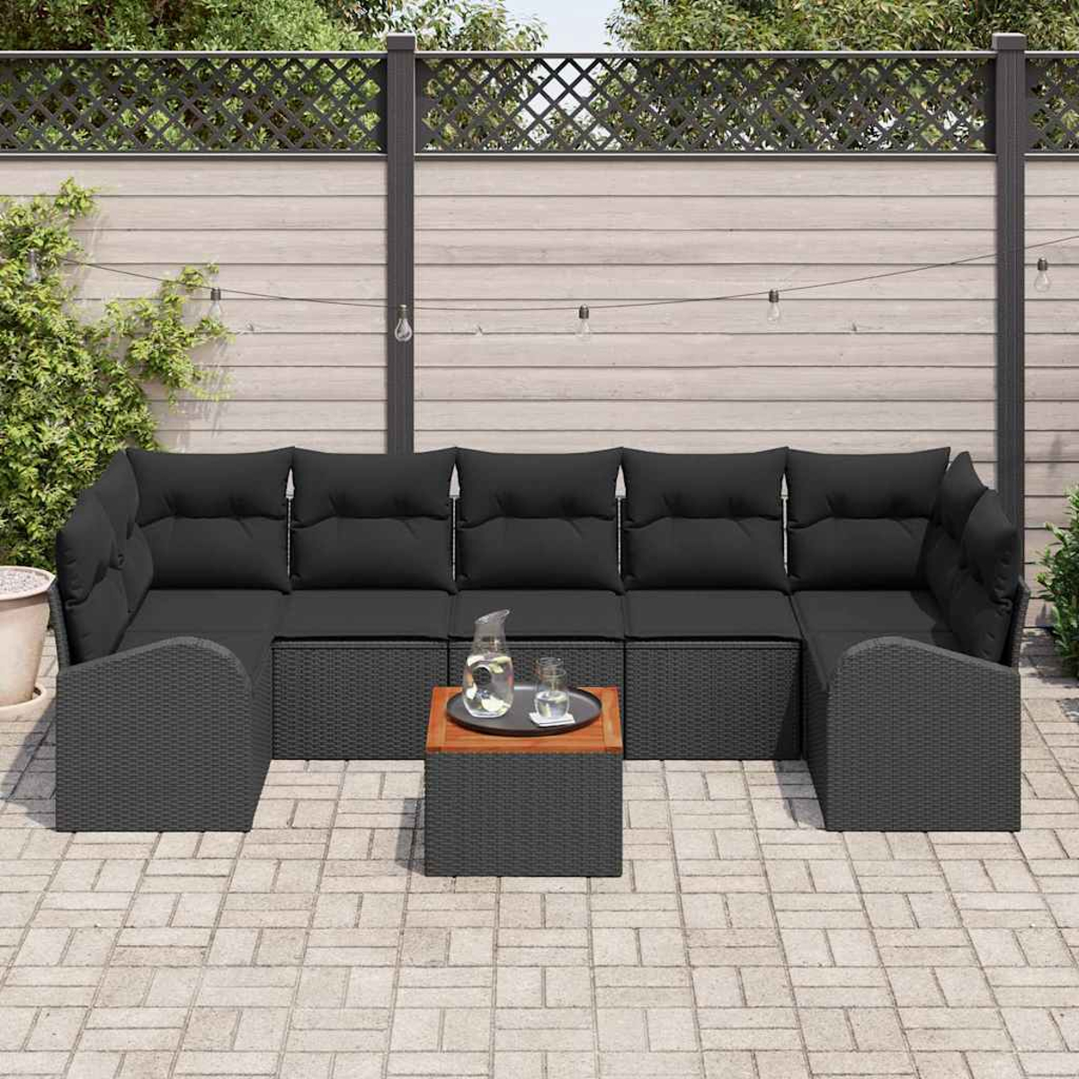 Gartensektionssofa mit schwarzen Kissen. Ein kleiner Tisch mit Tablett und Krug. Zaun und Pflanzen im Hintergrund.