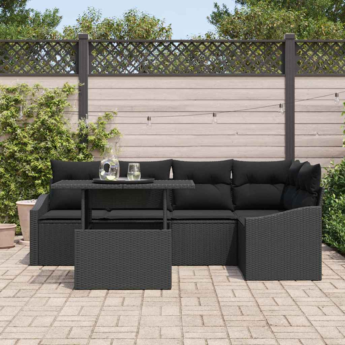 Schwarzes Outdoor-Geflechtmöbel-Set mit Kissen und Tisch. Im Hintergrund ein Zaun mit Pflanzen und eine Ziegelterrasse.