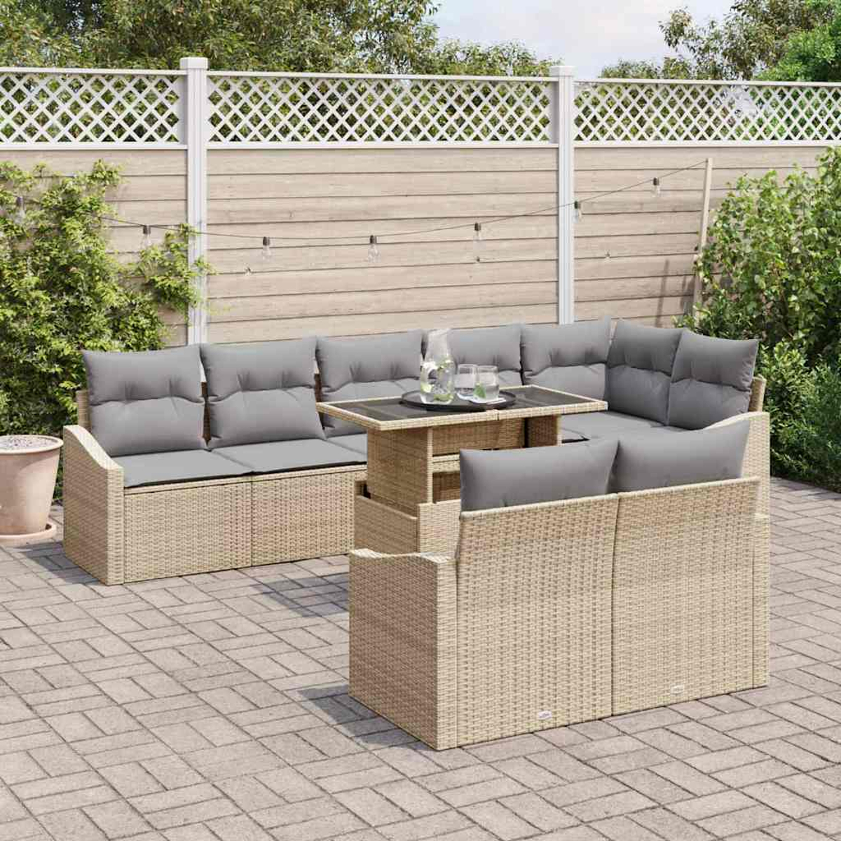 Outdoor-Korbschnitt mit grauen Kissen und einem Tisch mit Glasplatte auf einer Ziegelterrasse. Zaun und Grün im Hintergrund.