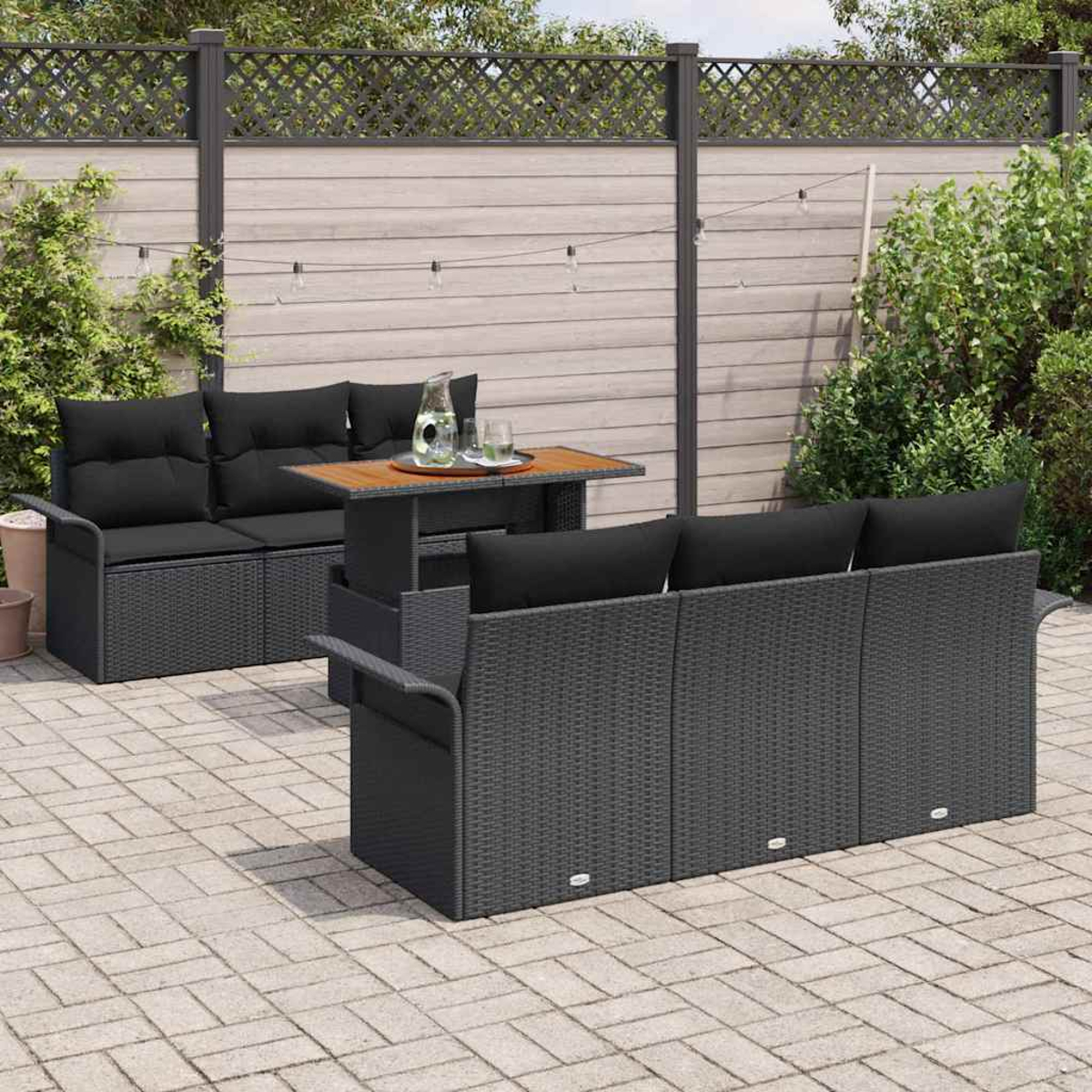 Outdoor-Terrassenset mit schwarzen Kissen und einem Holztisch auf einer Ziegelterrasse. Dahinter ein Holzzaun und grüne Büsche.