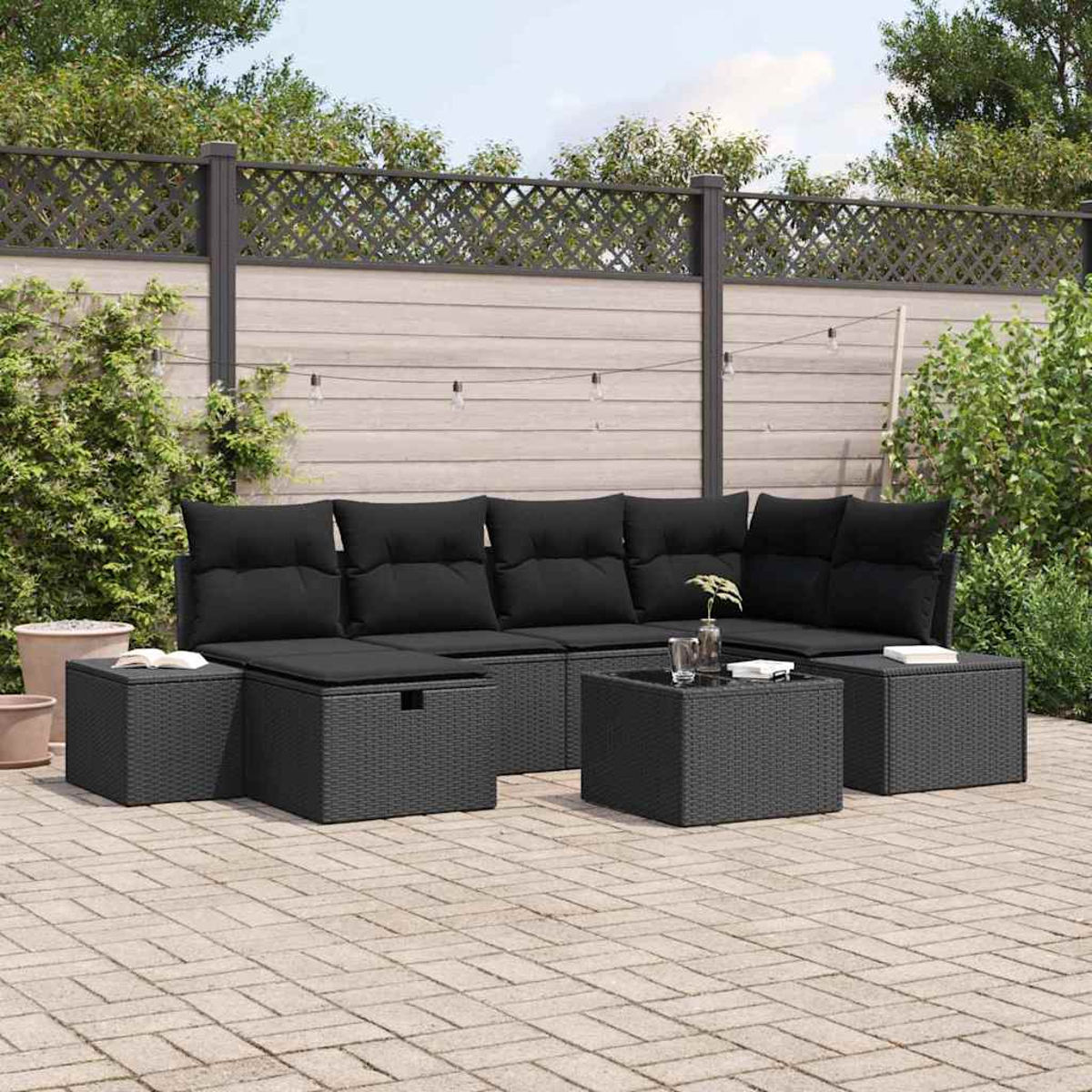 Terrassengarnitur mit schwarzen Kissen und gewebten Möbeln. Enthält ein Sofa, Ottomanen und einen Tisch. Vor einem Zaun und Grün.