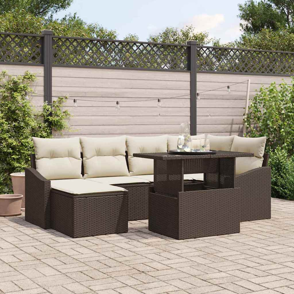 Terrassenset mit braunen Korbmöbeln, beigen Kissen und einem Hubtisch auf einer gepflasterten Terrasse mit Holzzaun im Hintergrund.