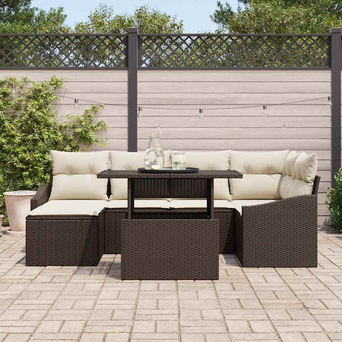 Garten-Terrassenset mit braunen Korbmöbeln, beigen Kissen und einem Tisch. Ein Zaun mit Lichtern und Pflanzen im Hintergrund.
