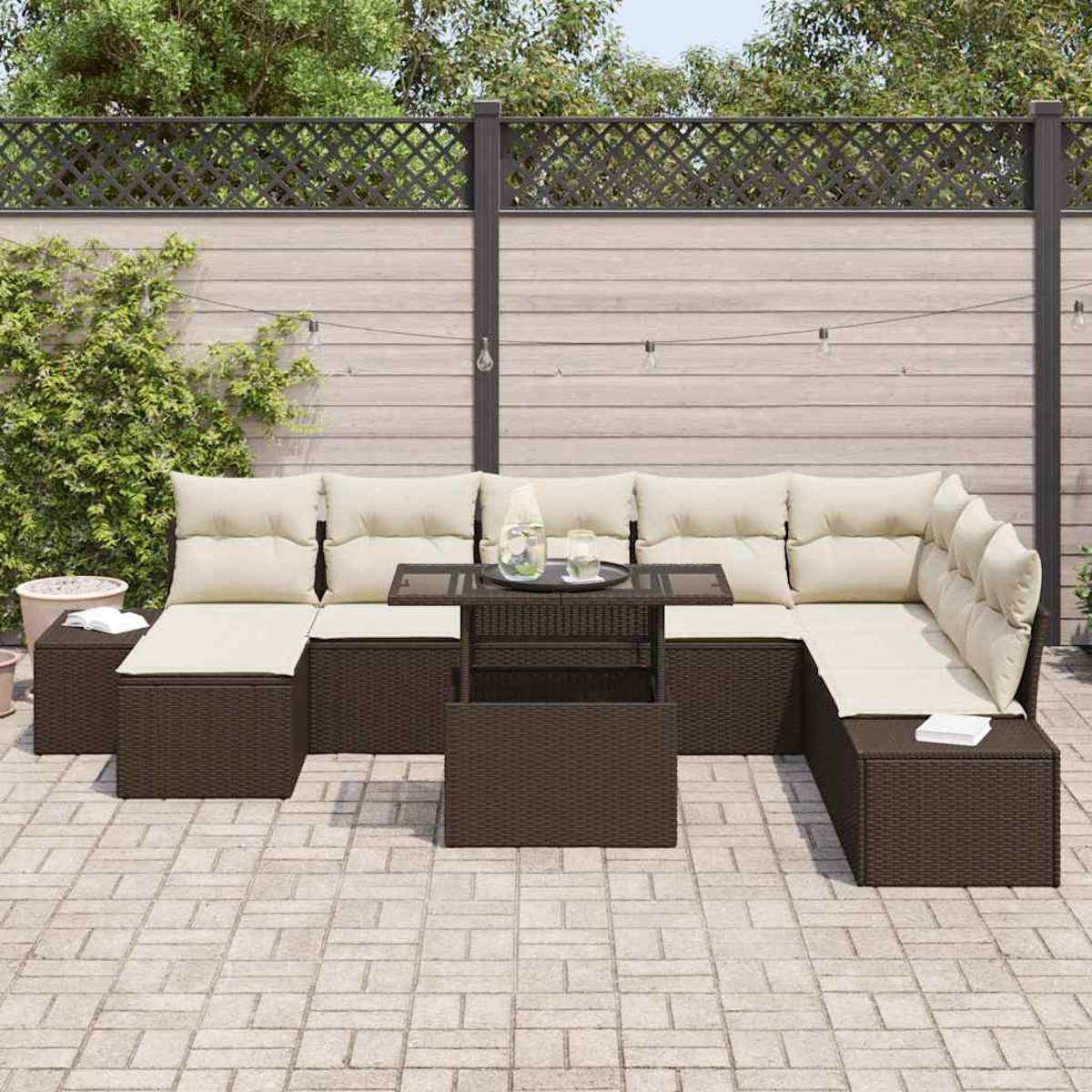 Garten-Terrassenset mit braunen Korbmöbeln, beigen Kissen und einem Couchtisch mit Glasplatte. Vor einem Holzzaun und Grün.