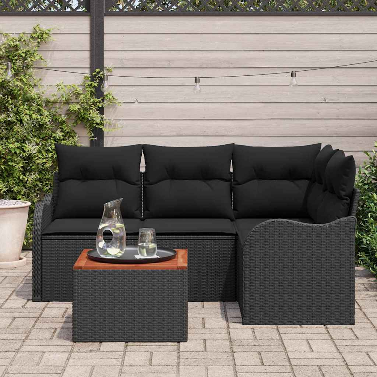 Terrassenset im Freien mit schwarzen Kissen. Davor steht ein kleiner Tisch mit Getränken. Grün und ein Holzzaun sind im Hintergrund.