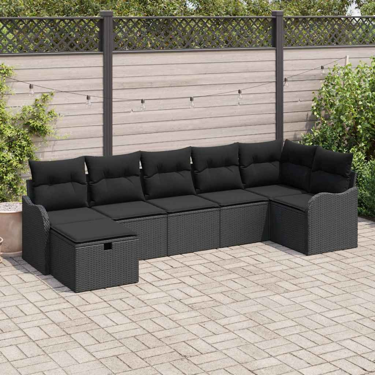 Un sofá seccional gris para exteriores con cojines negros en un patio de ladrillos. Una valla de madera y vegetación al fondo.
