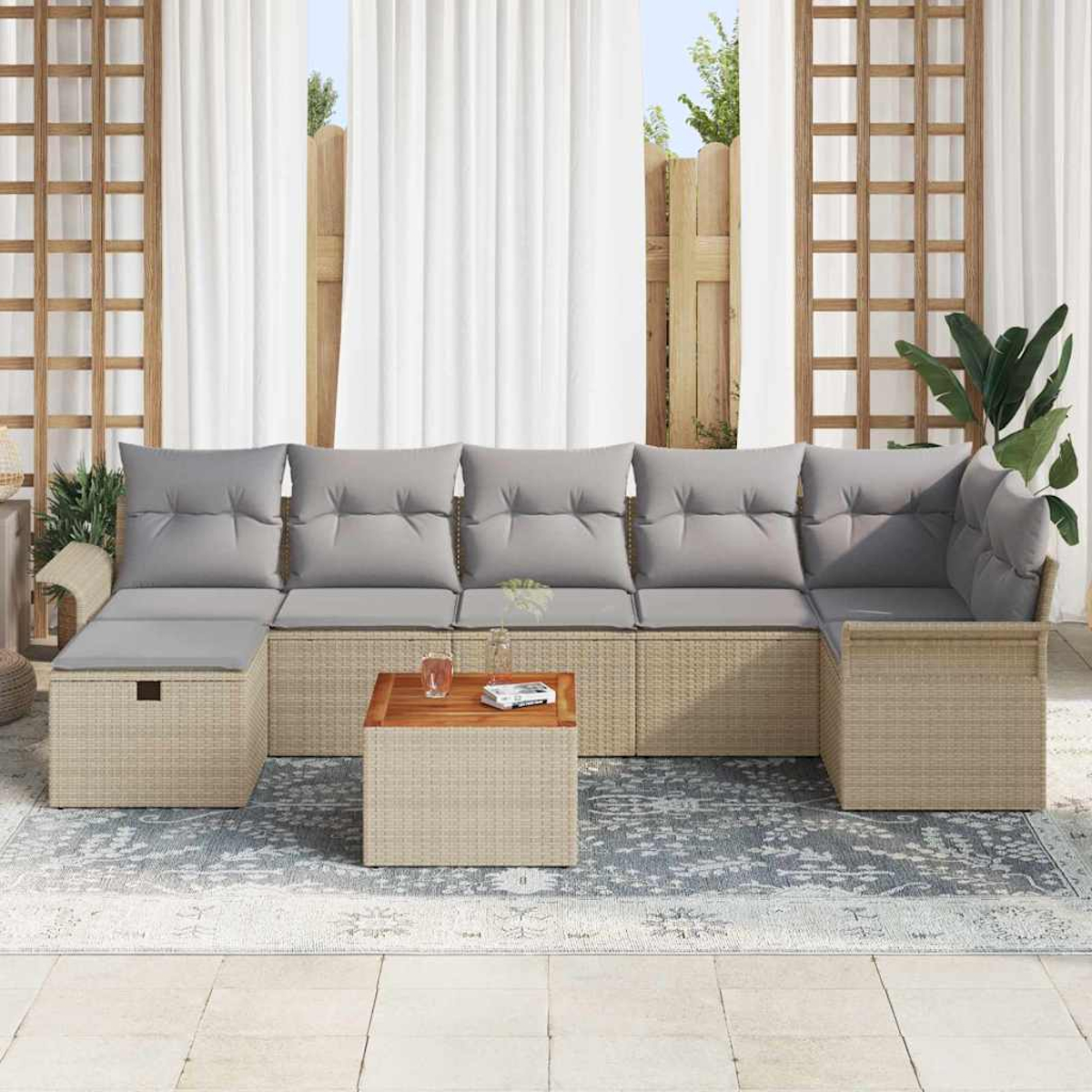 Outdoor-Sofa aus Korbgeflecht mit grauen Kissen und einem Holztisch auf einem gemusterten Teppich. Weiße Vorhänge im Hintergrund.
