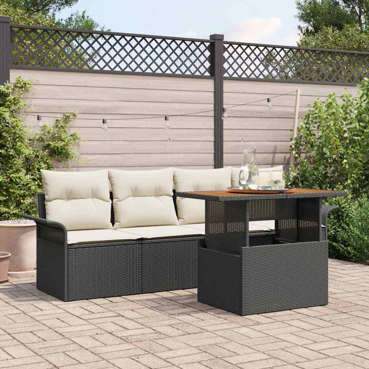 Outdoor-Terrassenset mit dunklem Korbmöbelsofa, Tisch und cremefarbenen Kissen. Holzzaun und Grün im Hintergrund. Lichterketten sind sichtbar.