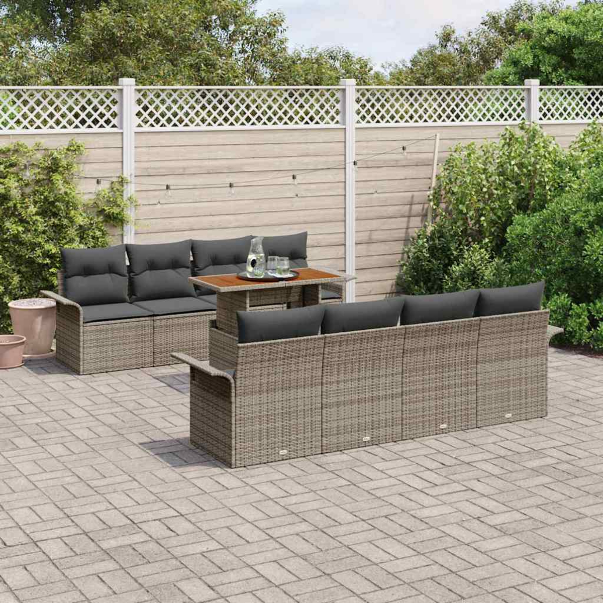 Gartenmöbel-Set. Rattan-Sofa, Tisch mit Glas und Flasche. Graue Kissen. Gepflasterte Terrasse, Holzzaun und grüne Büsche im Hintergrund.
