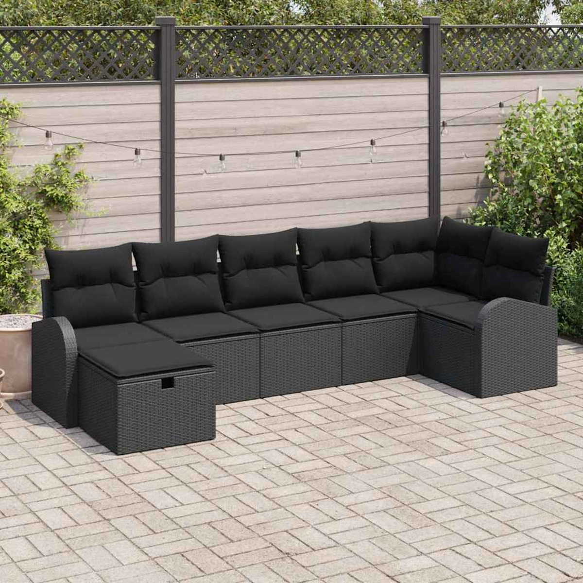 Outdoor-Ecksofa mit schwarzen Kissen. Graue Korbbasis. Auf einer gepflasterten Terrasse, mit Holzzaun und Grün im Hintergrund.