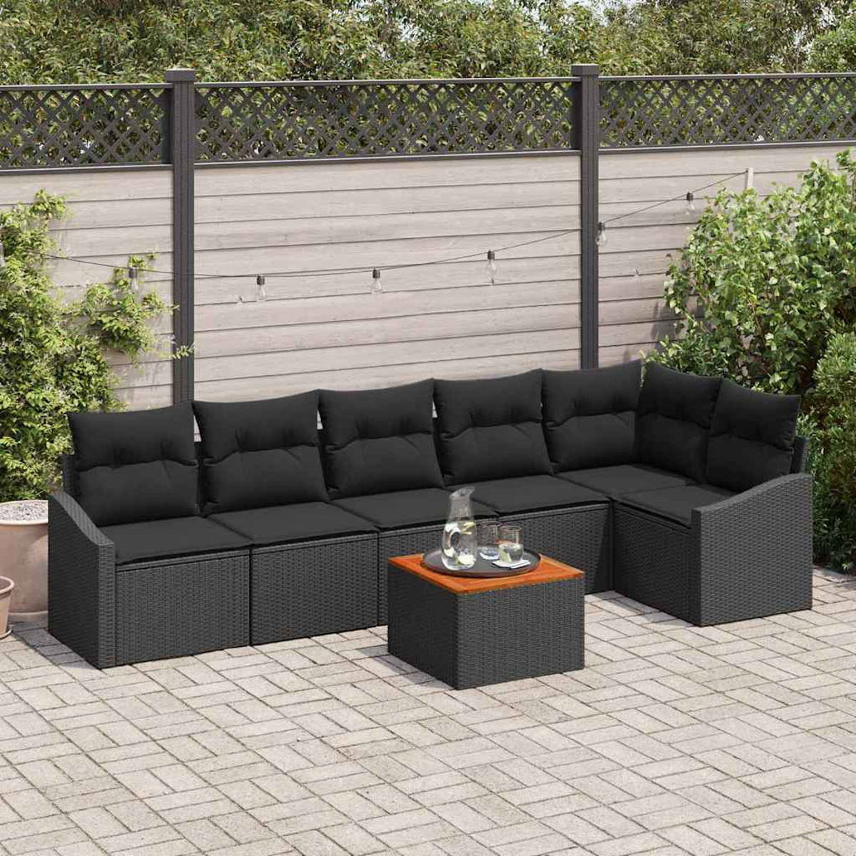 Escena al aire libre. Un sofá seccional negro de mimbre con cojines, una mesa cuadrada y una valla de madera al fondo. Una jarra y vasos están sobre la mesa.