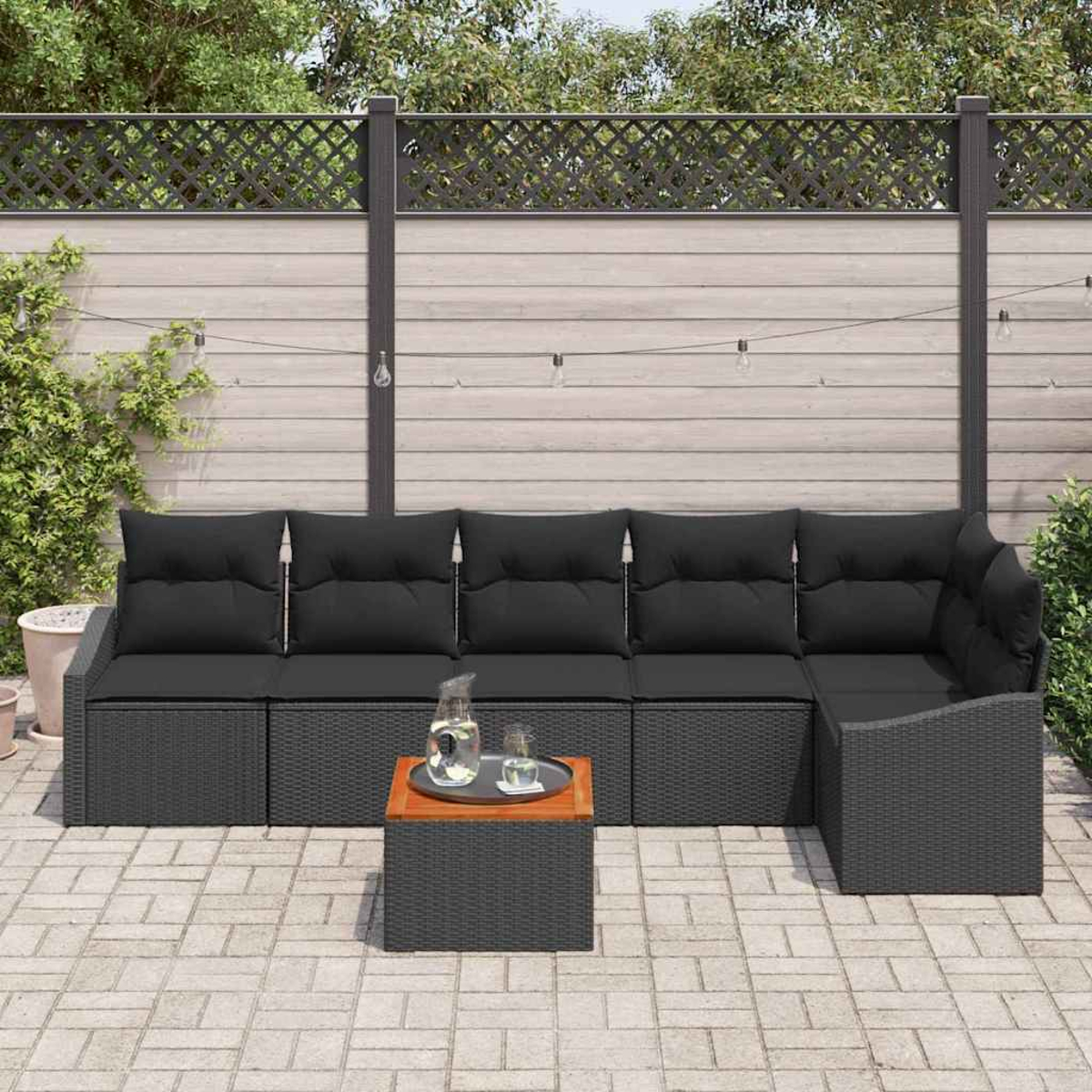 Sofá de mimbre negro para exteriores con cojines, mesa pequeña con bebidas, en un patio pavimentado. Valla de madera al fondo.