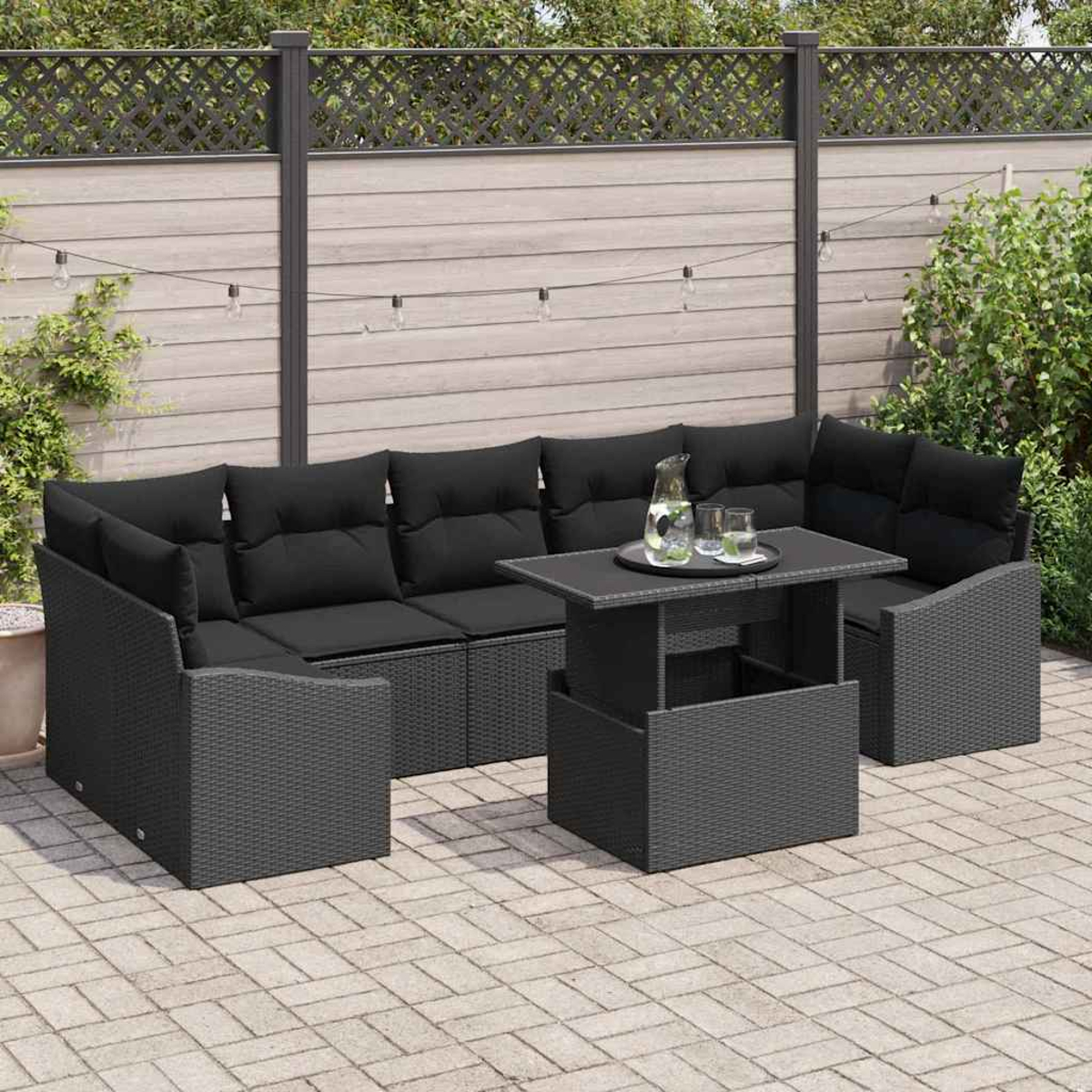Gartenmöbel-Set mit schwarzem Sofa und Tisch. Auf dem Tisch stehen eine Kanne und Gläser. Hintergrund: Holzzaun und Pflanzen.