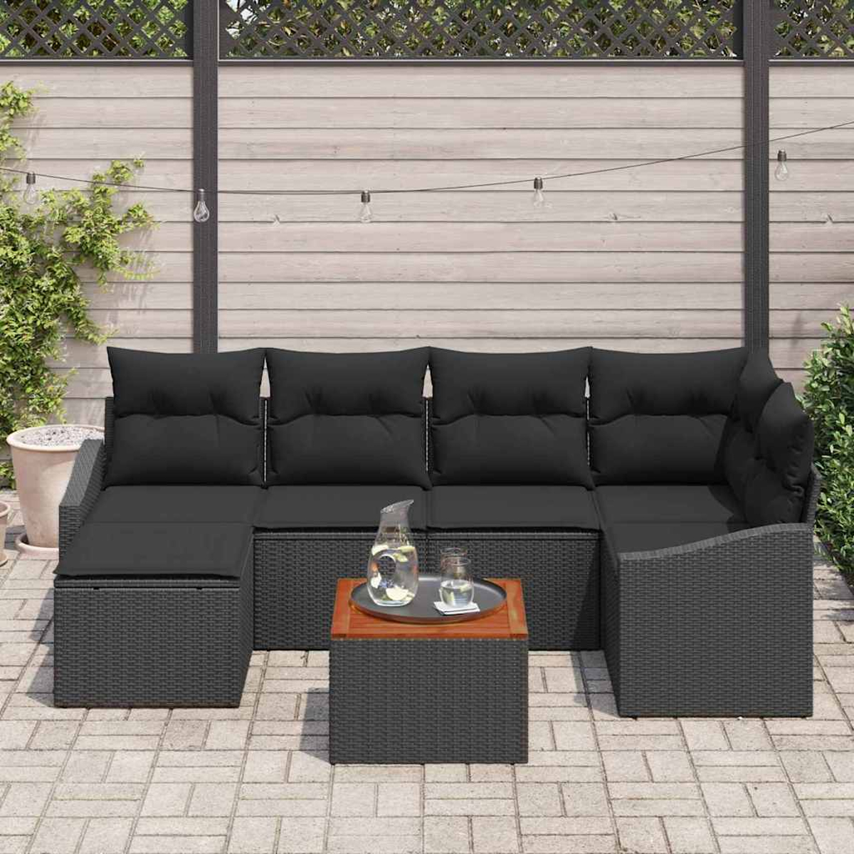 Gartenmöbel mit schwarzen Kissen. Ein kleiner Tisch mit Tablett, Krug und Gläsern. Hintergrund: Holzzaun.