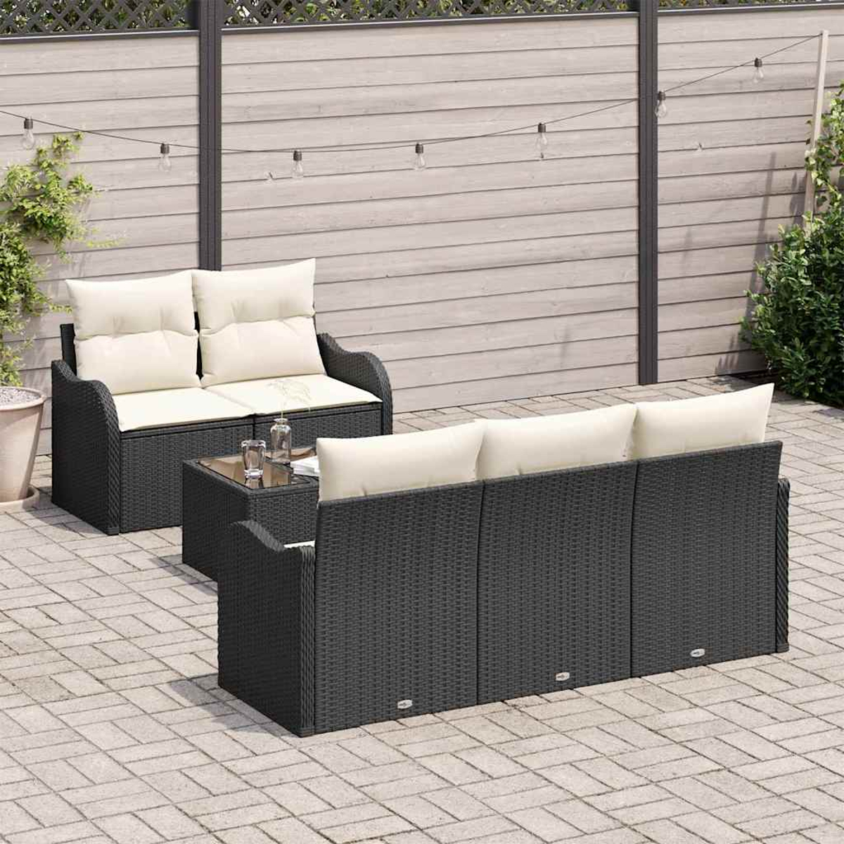 Gartenmöbel-Set. Schwarzes Korbmöbel-Sofa mit cremefarbenen Kissen, kleiner Tisch, Hintergrund Zaun und Grün auf der Terrasse.