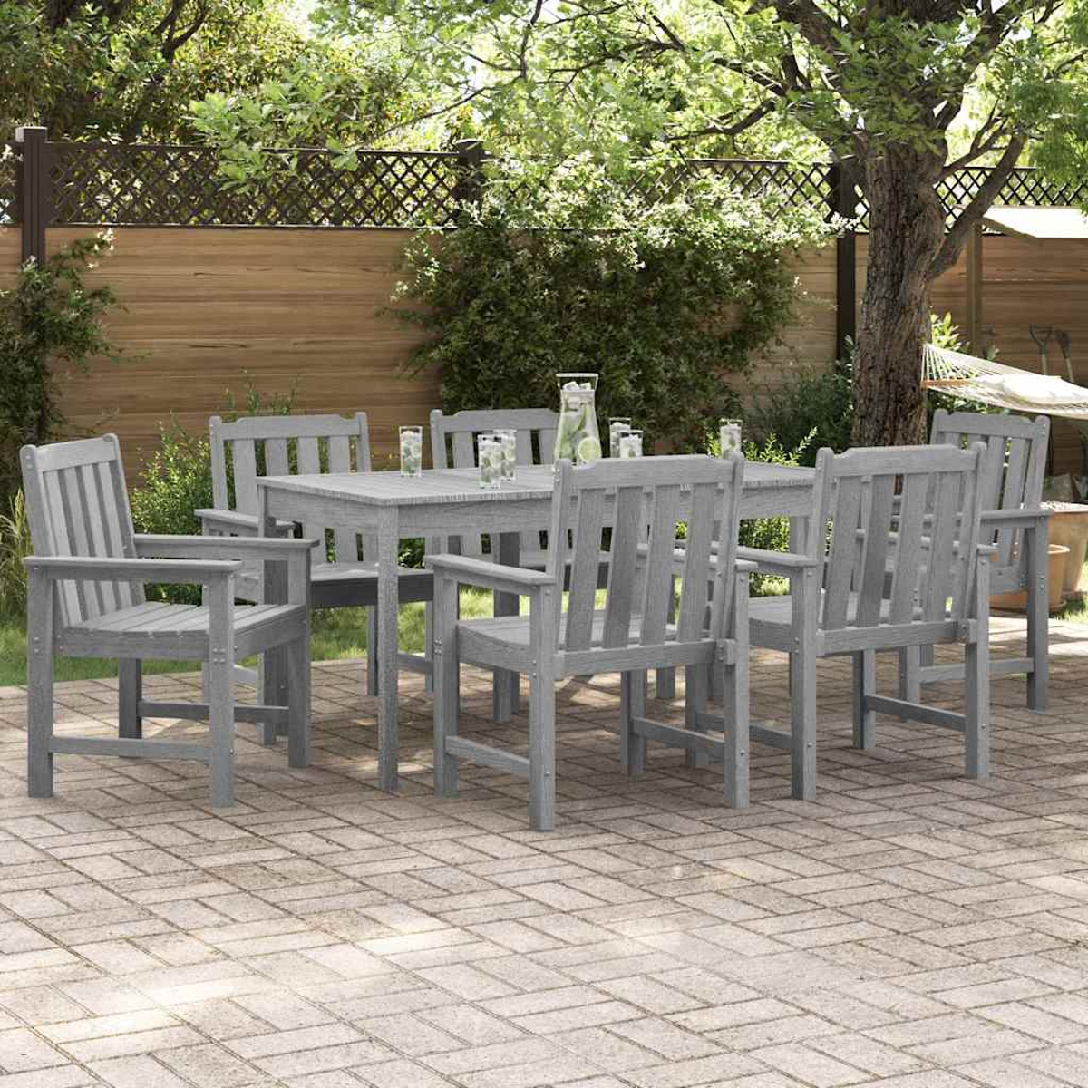 Ein graues Ess-Set aus Holz im Freien auf einer Backstein-Terrasse. Enthält einen Tisch, Stühle und Getränke. Grün und eine Hängematte im Hintergrund.