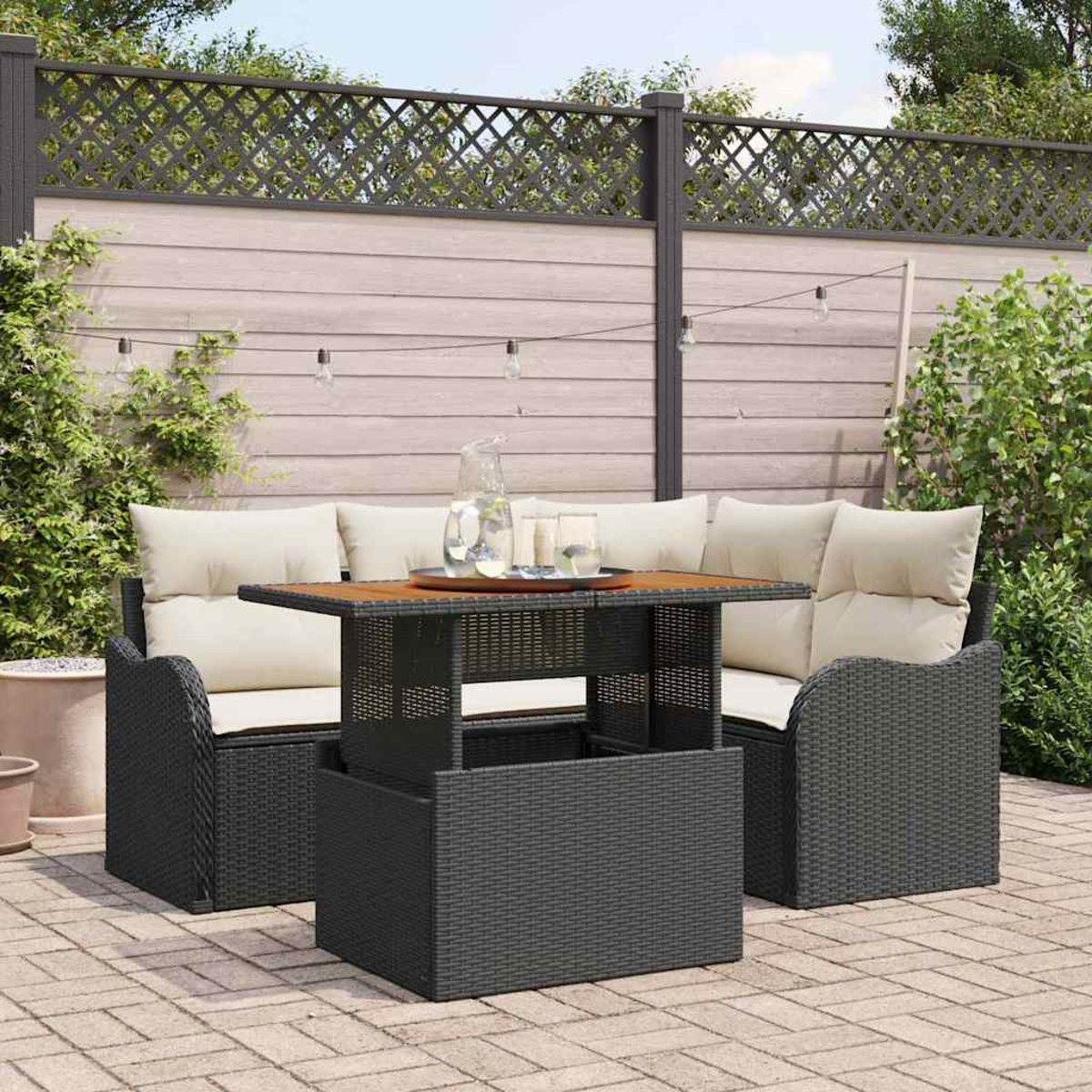 Juego de asientos al aire libre con mesa. Muebles de mimbre negro, cojines crema, tablero de madera. Fondo: valla, plantas y luces.