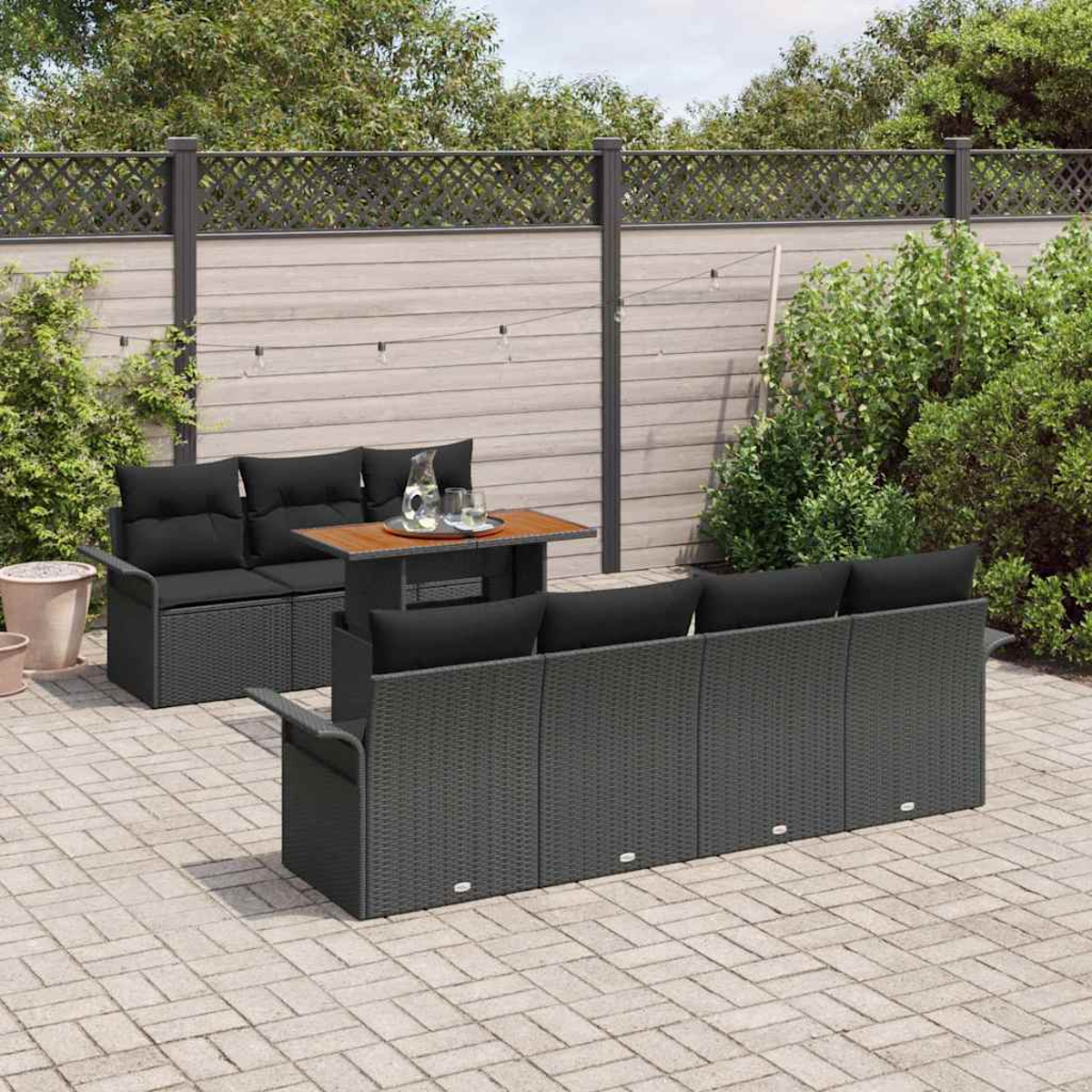 Juego de patio al aire libre con cojines negros y una mesa de madera. En un patio pavimentado, con una valla y vegetación de fondo.