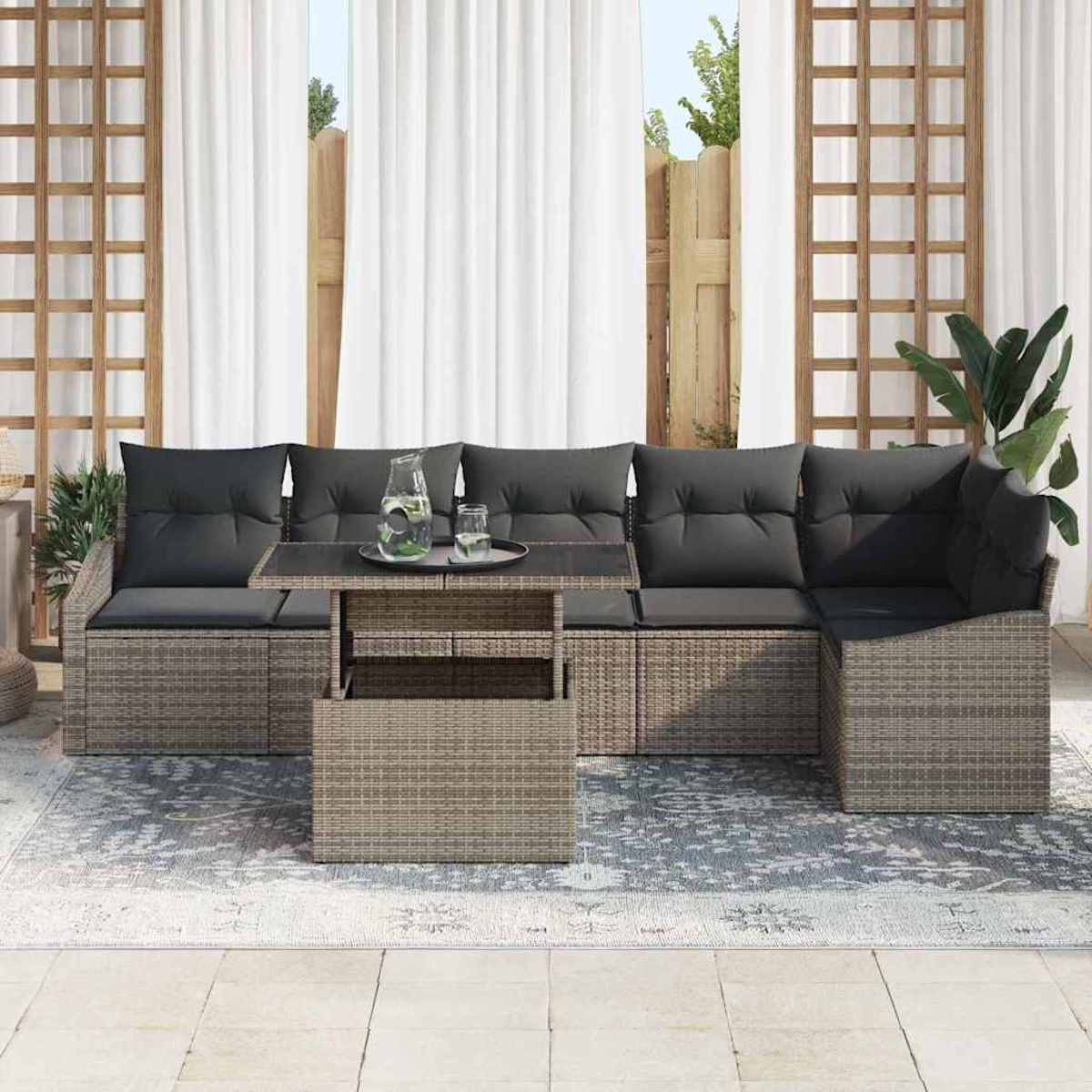 Outdoor-Ecksofa mit grauen Kissen, Glastisch mit Getränken, auf einem gemusterten Teppich. Weiße Vorhänge und Holzzaun im Hintergrund.