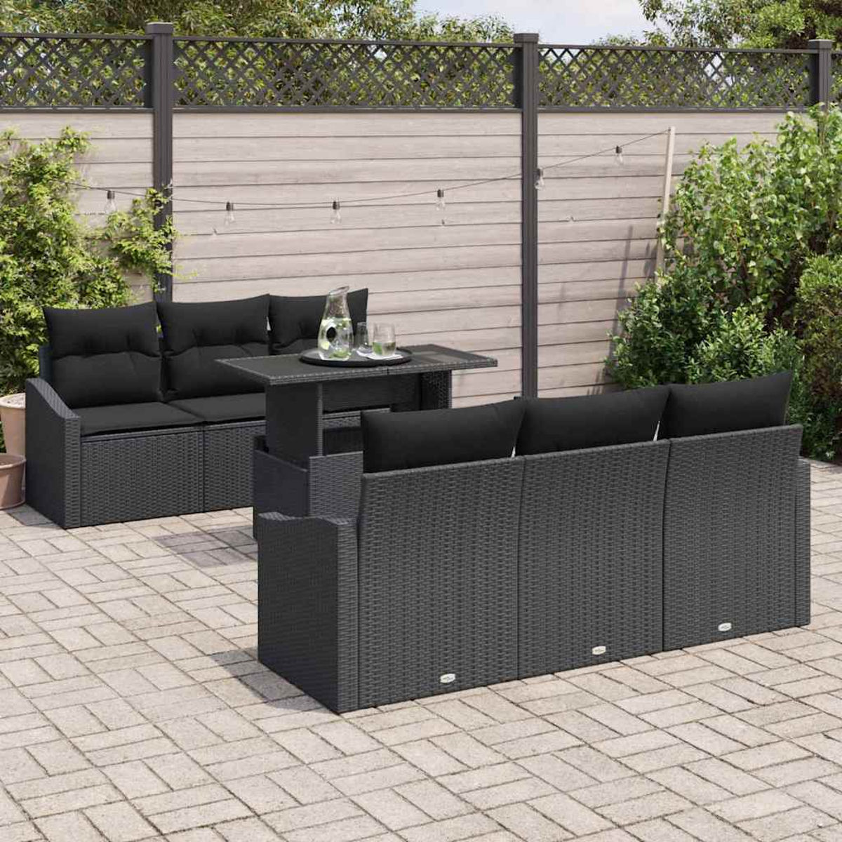 Garten-Terrassenset mit schwarzen Kissen, einem Tisch mit Getränken, auf einer Backstein-Terrasse. Ein Holzzaun und Grün im Hintergrund.