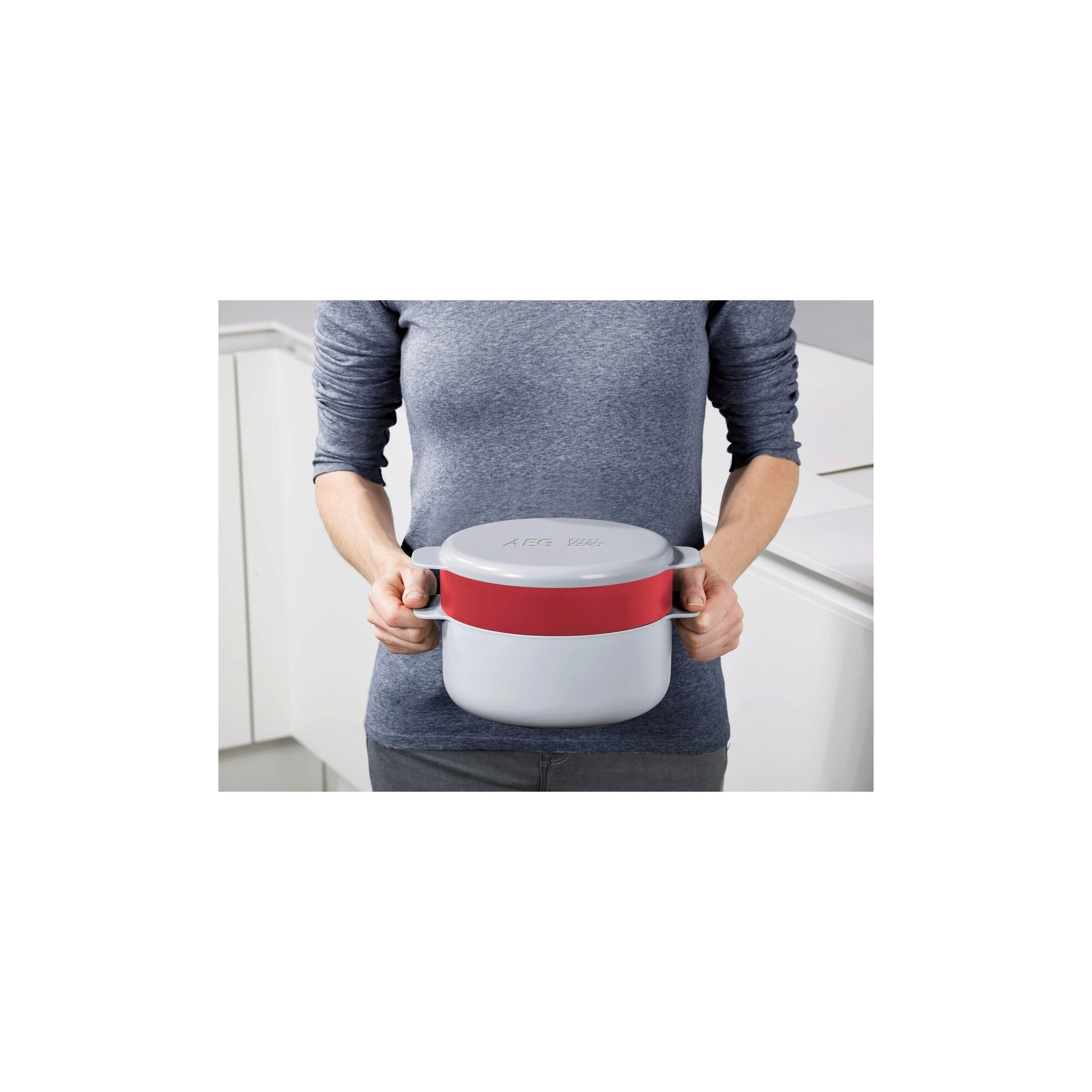 Una persona sostiene una vaporera AEG blanca y roja. La persona lleva una camisa gris y está en una cocina.