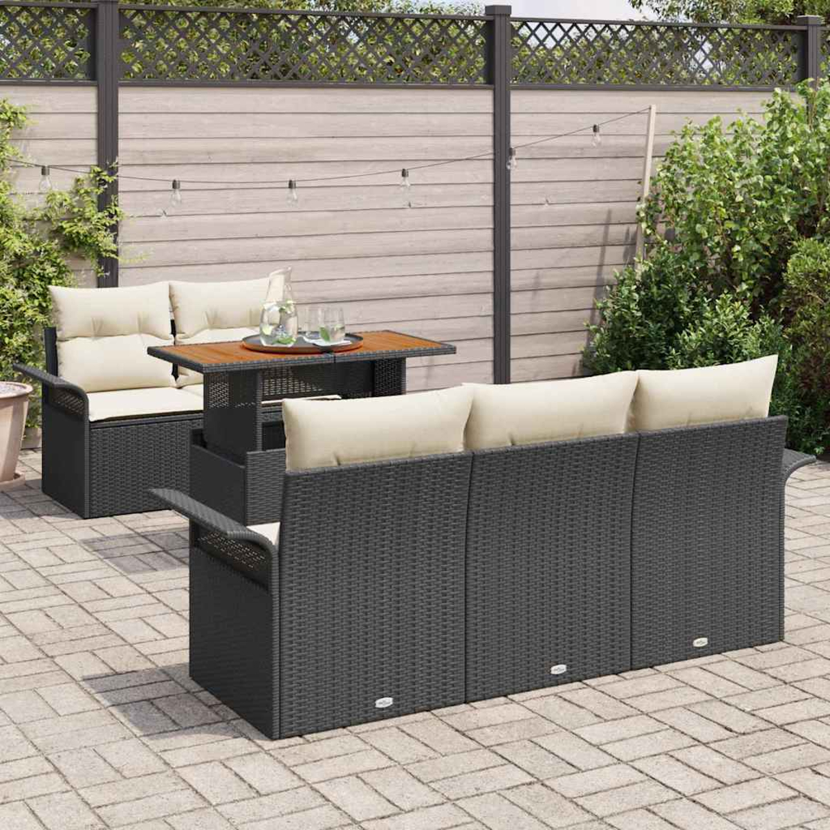 Garten-Terrassenset mit schwarzem Korbmöbel, cremefarbenen Kissen und einem Tisch. Ein Gartenzaun und Pflanzen im Hintergrund.