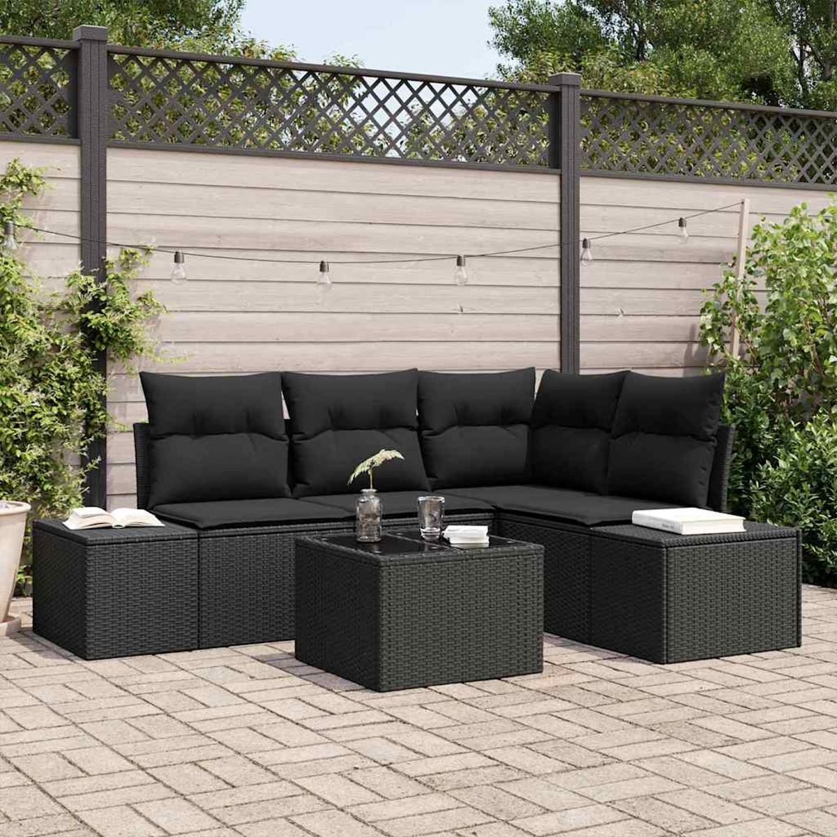 Ein Terrassenset mit schwarzen Kissen und einem Couchtisch. Ein Holzzaun und grüne Pflanzen bilden den Hintergrund. Eine Ziegelterrasse.