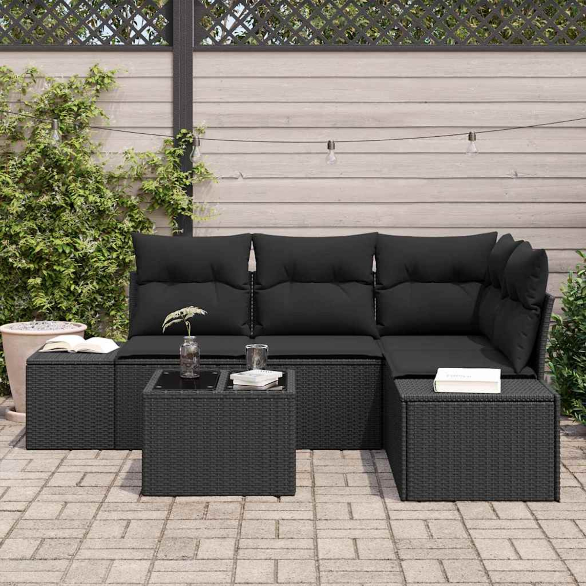 Schwarzes Outdoor-Sofa aus Rattan mit Kissen, einem Couchtisch und Ottomanen. Es steht auf einer gepflasterten Terrasse mit Zaun.