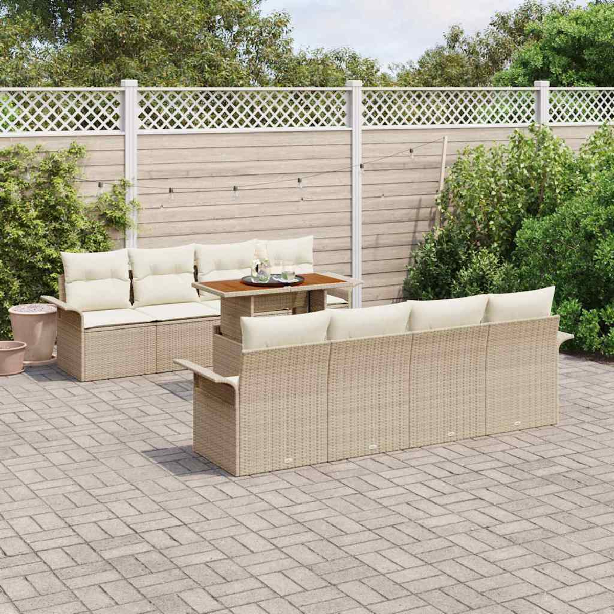 Terrassenset im Freien. Beige Korbsessel und Tisch mit cremefarbenen Kissen auf einer gepflasterten Terrasse. Holzzaun und Grün im Hintergrund.