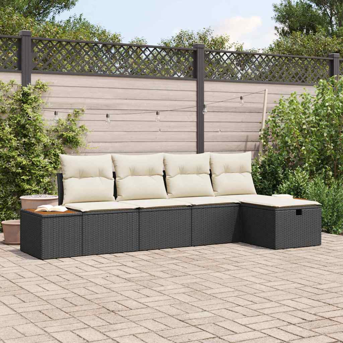 Ein schwarzes Outdoor-Sofa aus Korbgeflecht mit cremefarbenen Kissen steht auf einer Terrasse. Im Hintergrund befinden sich ein Holzzaun und Grün.