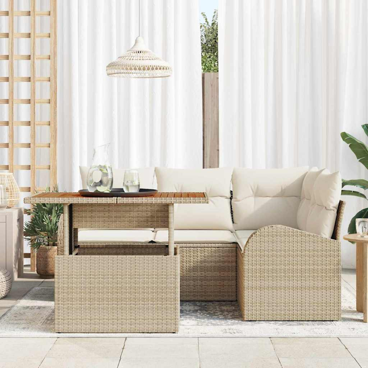 Zona de estar al aire libre con muebles de mimbre, una mesa de madera y cortinas blancas. Una jarra de agua y vasos en la mesa.