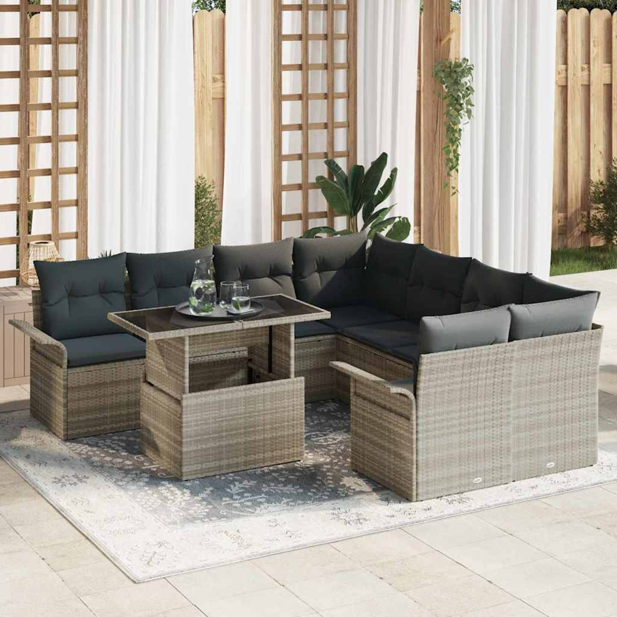 Outdoor-Rattansofa mit dunklen Kissen und einem Tisch. Auf einem gemusterten Teppich, in der Nähe eines weißen Vorhangs und Holzzauns.