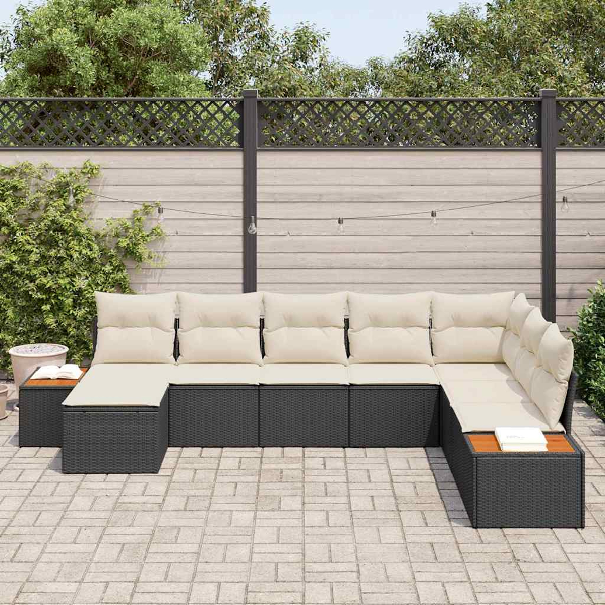 Ein Ecksofa im Freien mit beigen Kissen, schwarzem Rahmen und hölzernen Beistelltischen. Es steht auf einer Backstein-Terrasse, mit Zaun und Bäumen im Hintergrund.