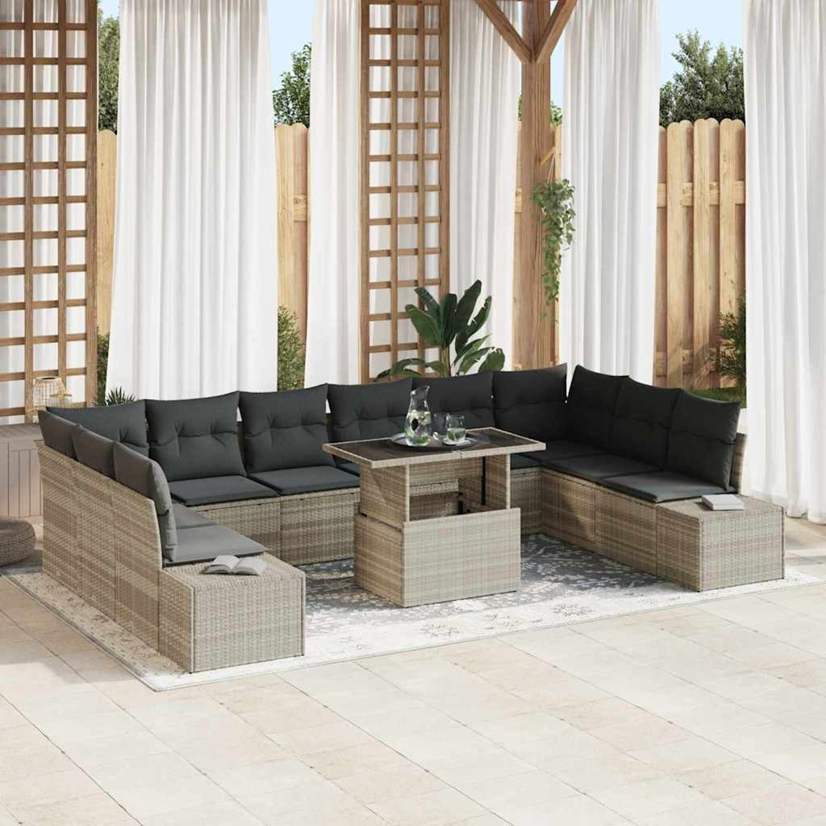 Outdoor-Sitzbereich mit Korbmöbeln, schwarzen Kissen und einem Tisch mit Hubfunktion. Weiße Vorhänge und Holzzaun im Hintergrund.